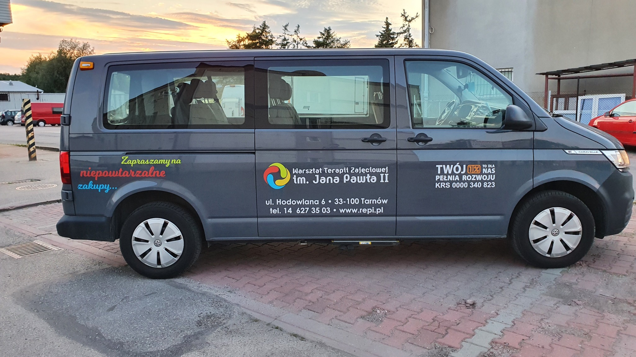 Szary samochód typu van z reklamowym oklejeniem dla Warsztatu Terapii Zajęciowej im. Jana Pawła II, z widocznym logo i danymi kontaktowymi.