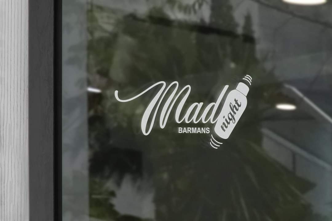 Biała naklejka z logo 'Mad Barmans night' umieszczona na szklanej witrynie lokalu, widoczne odbicie zieleni w szybie.
