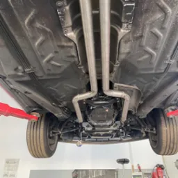 Widok podwozia samochodu na podnośniku, z nowym, podwójnym układem wydechowym ze stali nierdzewnej, mocowanym za pomocą metalowych obejm, widoczne elementy zawieszenia i skrzynia biegów.