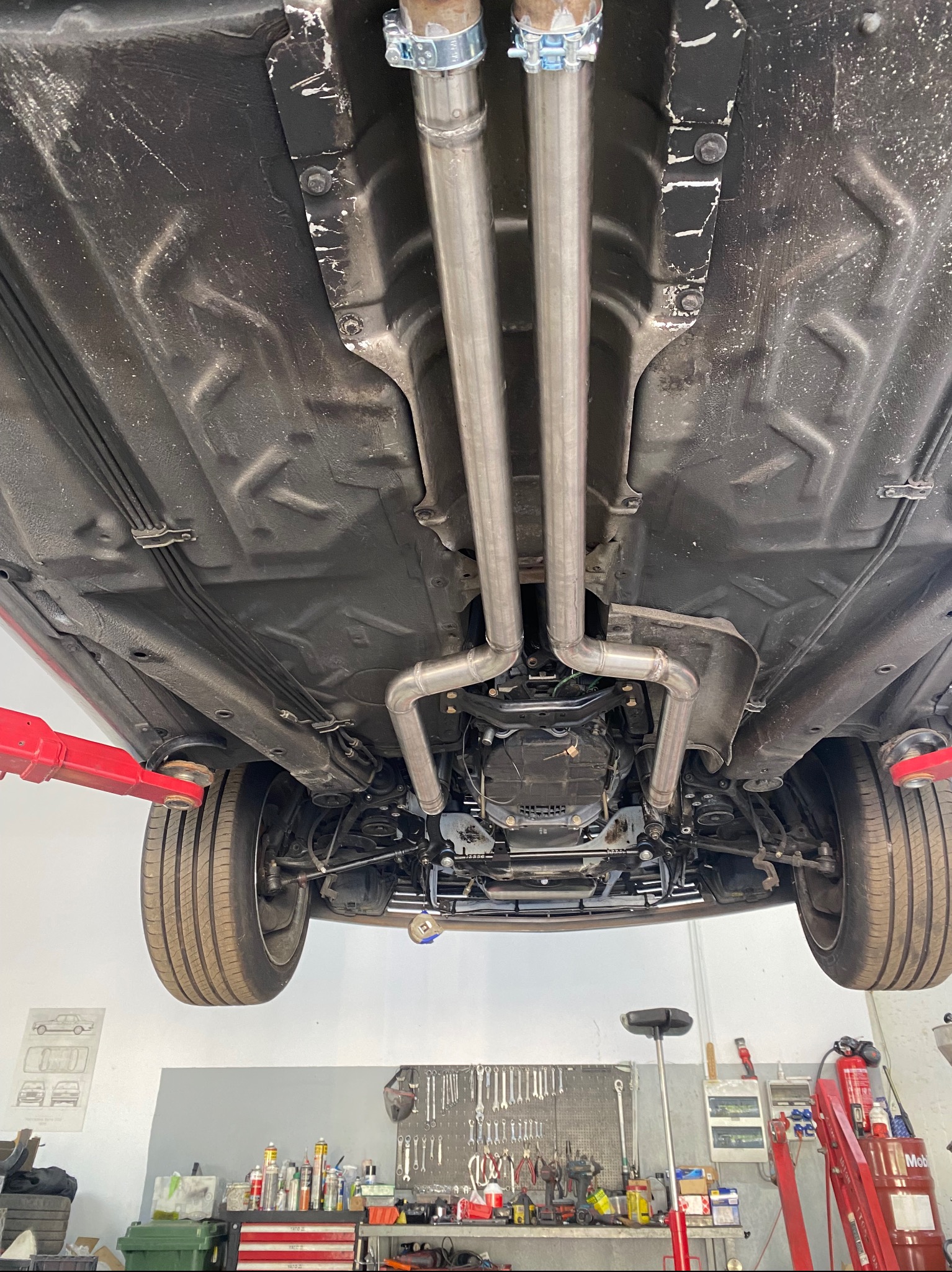 Widok podwozia samochodu na podnośniku, z nowym, podwójnym układem wydechowym ze stali nierdzewnej, mocowanym za pomocą metalowych obejm, widoczne elementy zawieszenia i skrzynia biegów.
