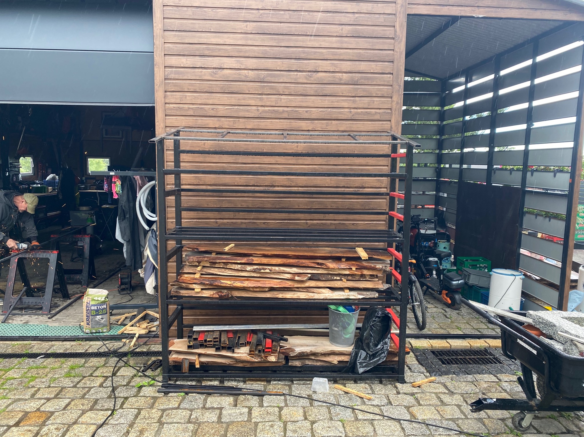 Warsztatowa scena z metalowym regałem wypełnionym drewnianymi elementami, w tle osoba obrabiająca metal z iskrami, widoczny materiał budowlany i taczka z kamieniami na brukowanym podwórku.