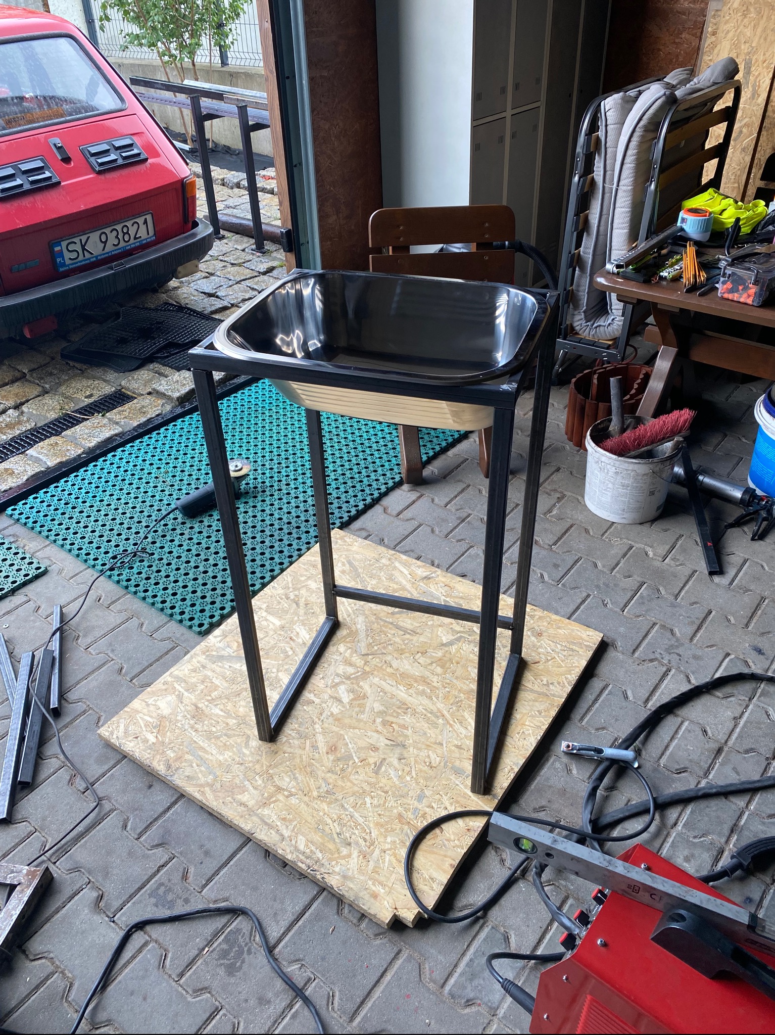 Metalowy stelaż z czarną misą na drewnianym podeście, widoczna spawarka, szlifierka kątowa i czerwony Fiat 126p w tle garażu.