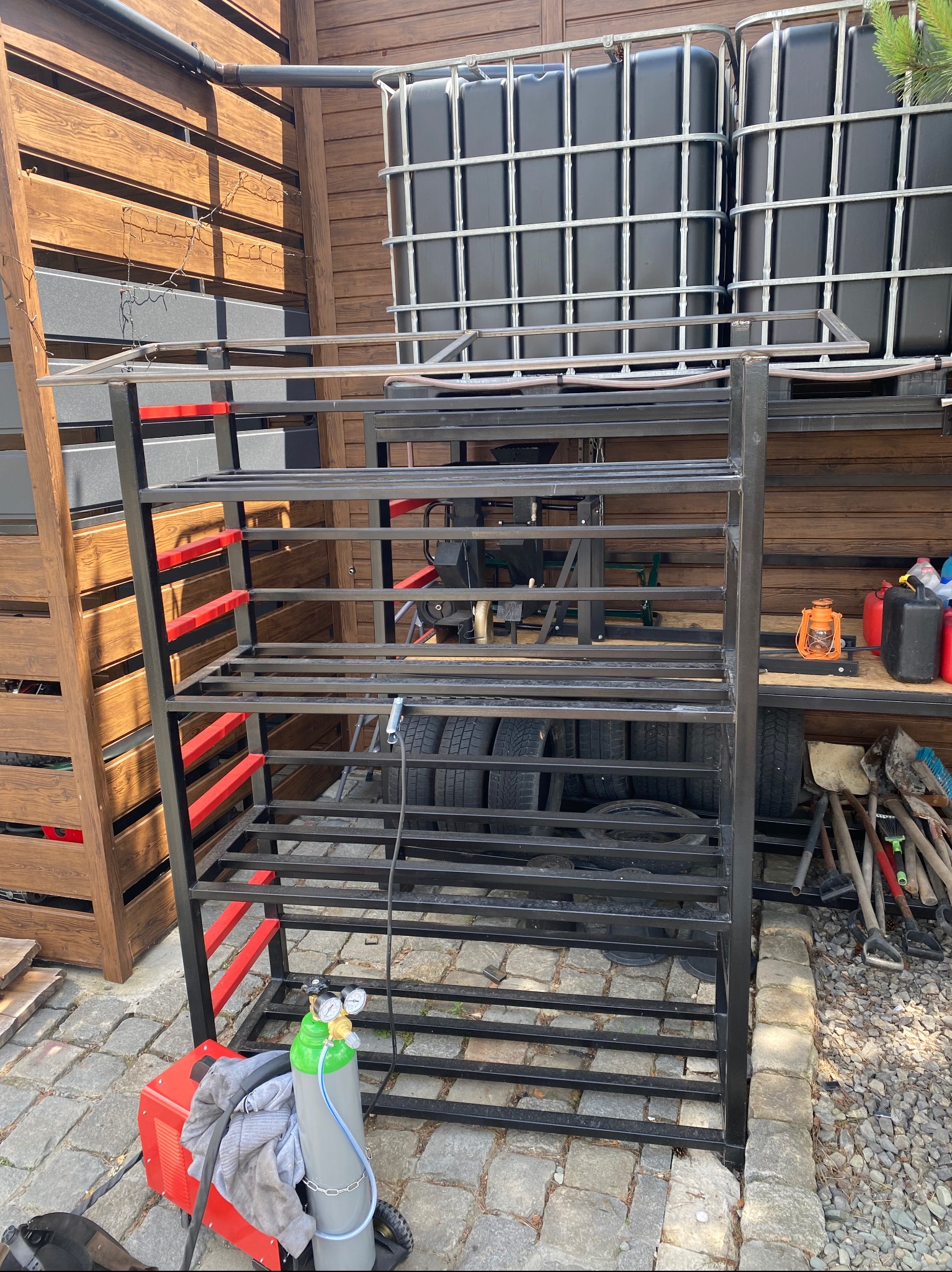 Metalowa konstrukcja regału z elementami pomalowanymi na czerwono, widoczny sprzęt spawalniczy, butla gazowa i narzędzia ogrodnicze w tle.