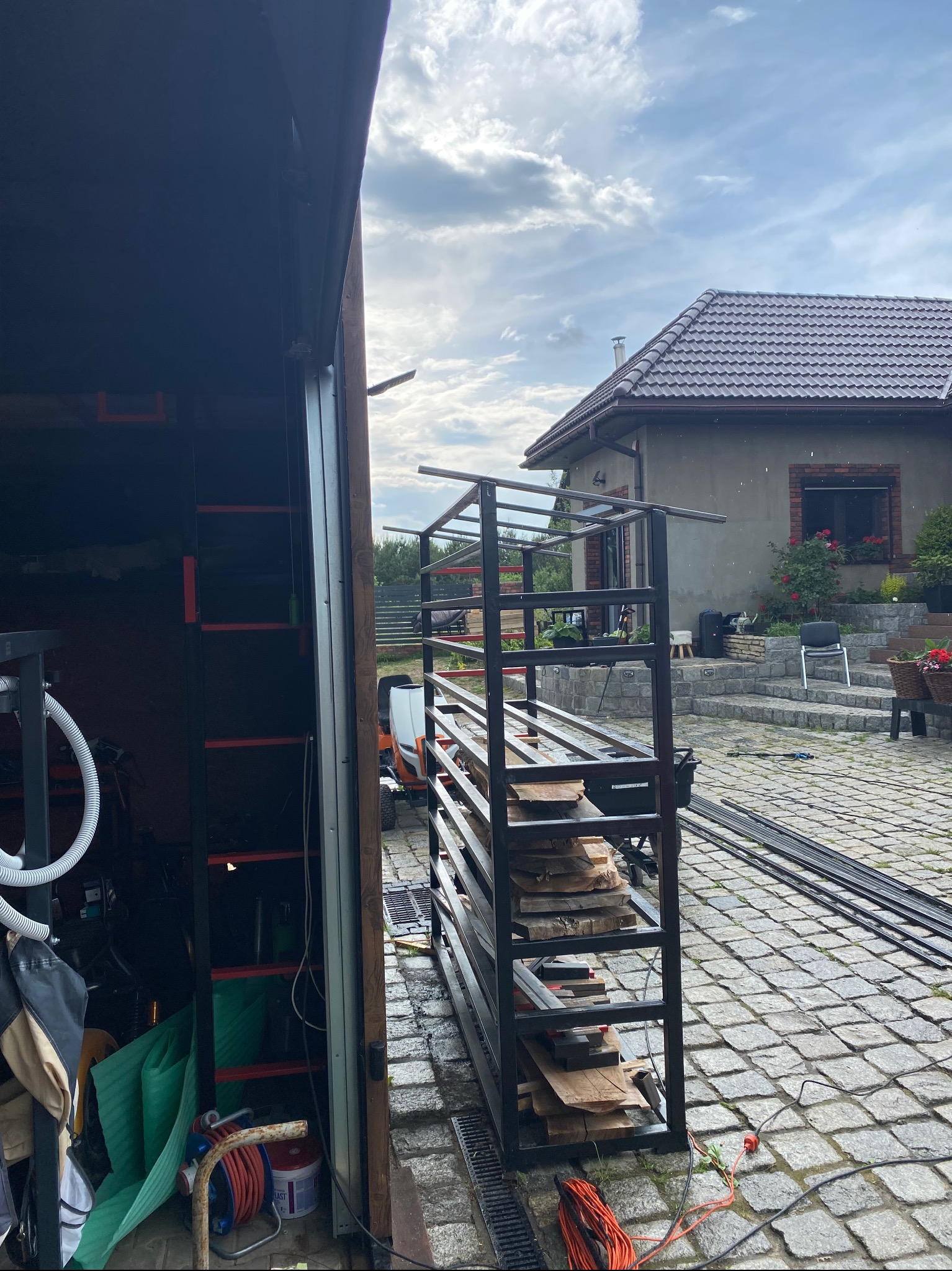 Konstrukcja metalowa z drewnianymi półkami, prawdopodobnie w trakcie montażu, widoczna na tle domu z ogrodem i otwartego garażu.