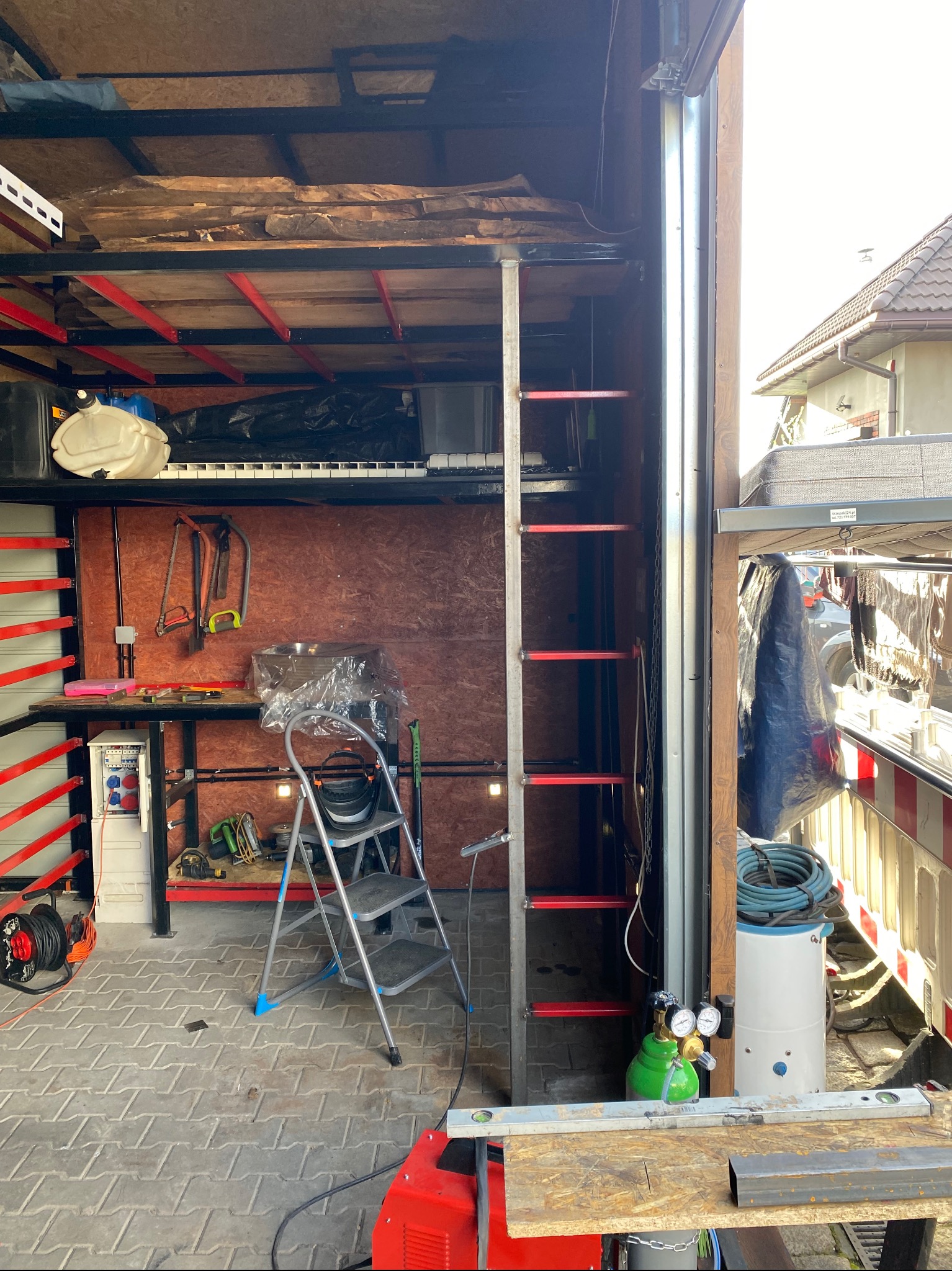 Wnętrze warsztatu z konstrukcją metalową, widoczne narzędzia, drabina aluminiowa, butla z gazem, spawarka i poziomica na stole roboczym, na górnej półce pianino i pojemniki.