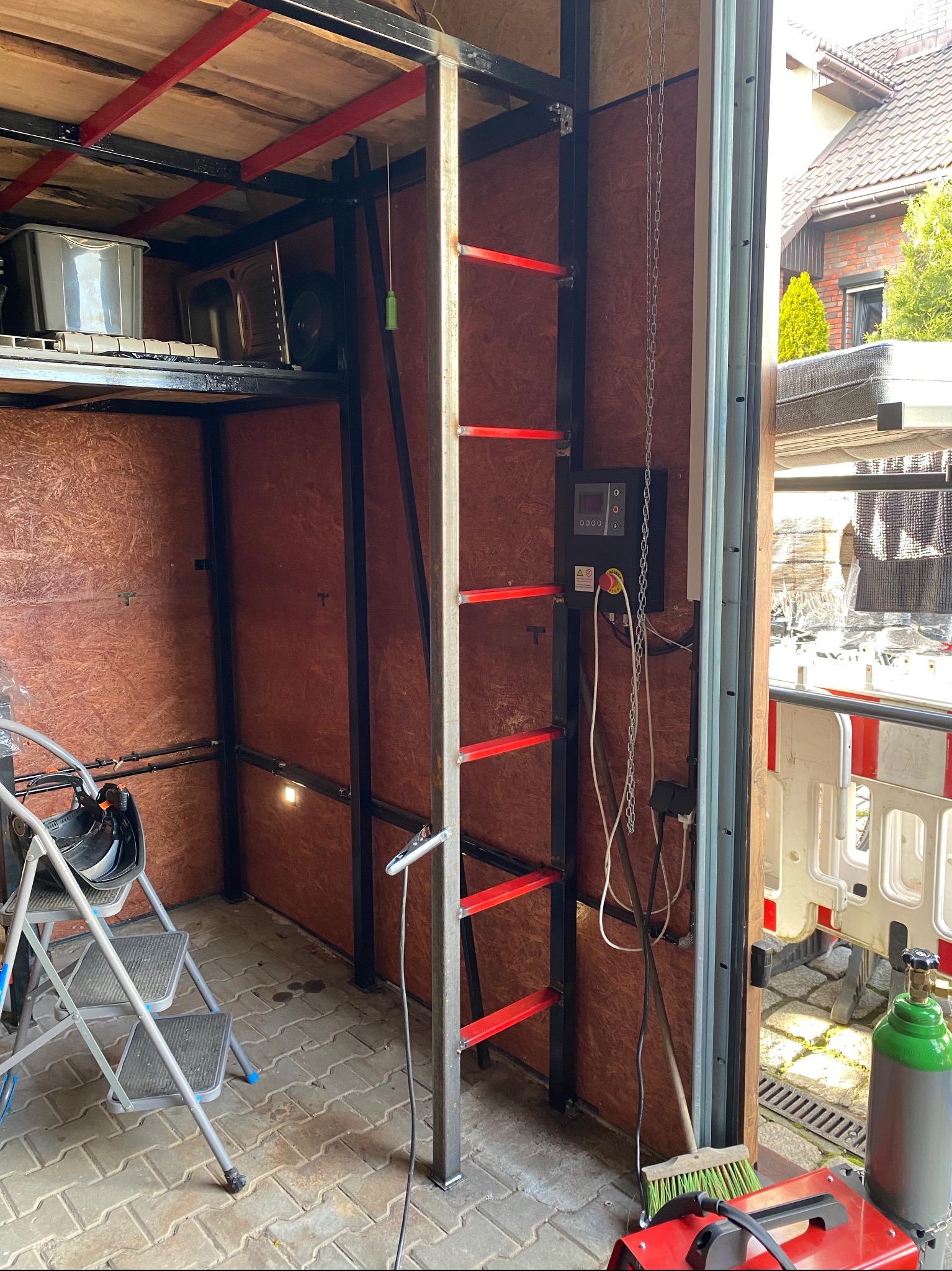 Wnętrze warsztatu z metalową konstrukcją, drabina z czerwonymi szczeblami, spawarka, butla z gazem i schodki.