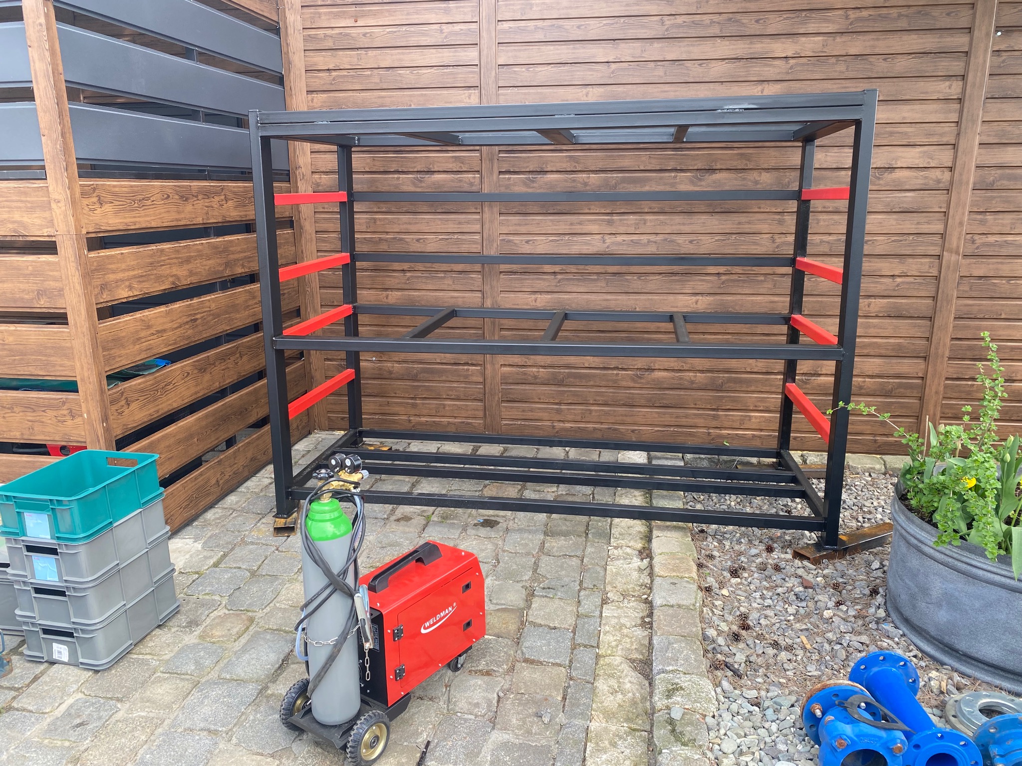 Metalowa konstrukcja regału z czerwonymi poprzeczkami, częściowo spawana, stoi na kostce brukowej obok spawarki, butli z gazem i stosu plastikowych pojemników.