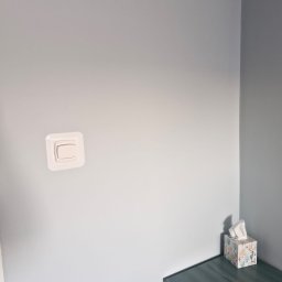 KISZNIEWSKI PAWEŁ KISZNIEWSKI - Świeżo pomalowana, jasnoniebieska ściana z zamontowanym białym włącznikiem światła i małym stolikiem z pudełkiem chusteczek w rogu pokoju.