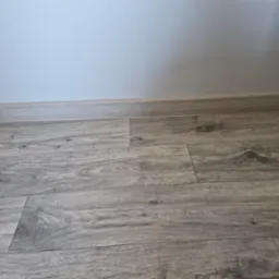 Szare panele podłogowe z drewnopodobnym wzorem ułożone wzdłuż ściany pomalowanej na jasny kolor, widoczna listwa przypodłogowa.