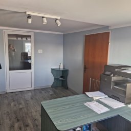 KISZNIEWSKI PAWEŁ KISZNIEWSKI - Biuro z biurkiem o zielonym blacie, drukarką, drewnianą podłogą, oświetleniem sufitowym i odzieżą wiszącą na wieszaku przy drzwiach.