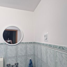 KISZNIEWSKI PAWEŁ KISZNIEWSKI - Biała łazienka z okrągłym lustrem, w którym odbija się osoba robiąca zdjęcie telefonem, nad umywalką i baterią oraz dekoracyjną listwą nad płytkami.