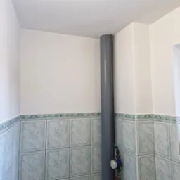 Szary pion kanalizacyjny w rogu łazienki z wykończonymi płytkami w tonacji błękitno-zielonej, widoczny manometr na rurze i fragment okna z rolkami papieru toaletowego na parapecie.