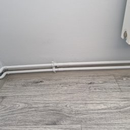 KISZNIEWSKI PAWEŁ KISZNIEWSKI - Dwie białe rury centralnego ogrzewania, pomalowane na biało, biegnące wzdłuż szarej podłogi panelowej i przy białej ścianie, widoczne obejmy mocujące.