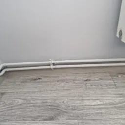 Dwie białe rury centralnego ogrzewania, pomalowane na biało, biegnące wzdłuż szarej podłogi panelowej i przy białej ścianie, widoczne obejmy mocujące.