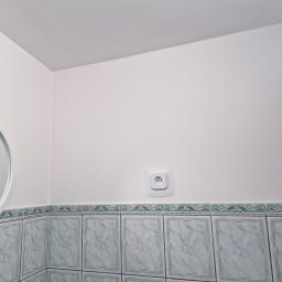 KISZNIEWSKI PAWEŁ KISZNIEWSKI - Biała ściana w łazience w Borzytuchom po malowaniu, z okrągłym lustrem i gniazdkiem elektrycznym nad pasem dekoracyjnych, marmurkowych płytek.