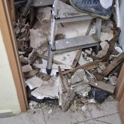 KISZNIEWSKI PAWEŁ KISZNIEWSKI - Demontaż podłogi w łazience: widoczne gruz, fragmenty płytek, drewniane elementy i drabina z butem na jednym ze stopni, przygotowanie do remontu.