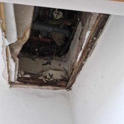 KISZNIEWSKI PAWEŁ KISZNIEWSKI - Otwór w suficie odsłaniający rury instalacyjne, widoczne uszkodzenia płyty kartonowo-gipsowej i drewnianej konstrukcji, fragment ściany z gniazdkiem elektrycznym w tle.