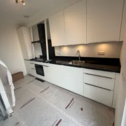 Nowoczesna kuchnia z białymi szafkami i czarnym blatem, oświetlona punktowo. Podłoga zabezpieczona folią ochronną. Minimalistyczny design.