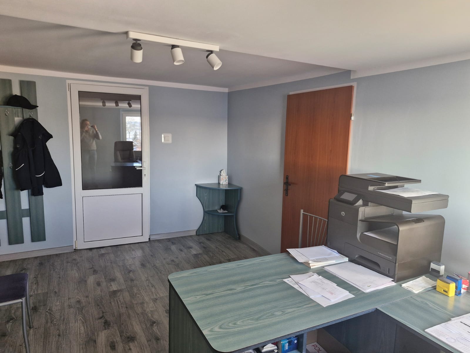 Biuro z biurkiem o zielonym blacie, drukarką, drewnianą podłogą, oświetleniem sufitowym i odzieżą wiszącą na wieszaku przy drzwiach.