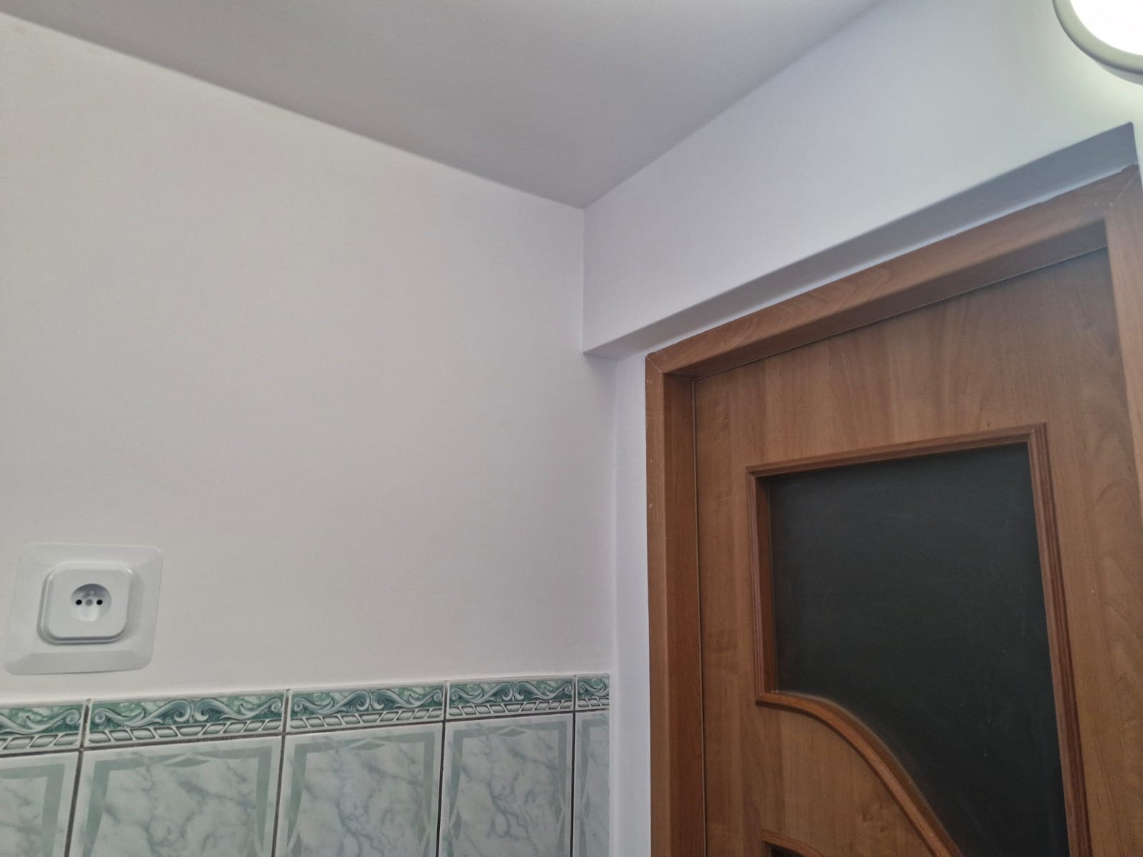 Fragment łazienki po remoncie: białe ściany, drewniane drzwi z ciemnym szkłem, ozdobne płytki ceramiczne i gniazdko elektryczne.