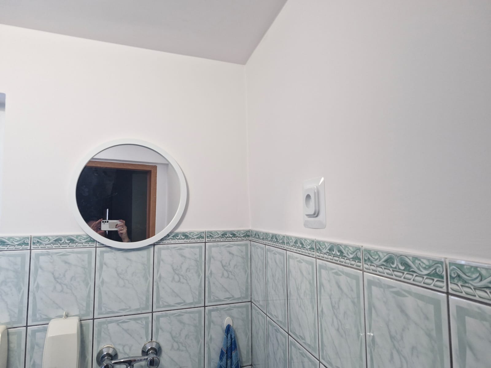 Biała łazienka z okrągłym lustrem, w którym odbija się osoba robiąca zdjęcie telefonem, nad umywalką i baterią oraz dekoracyjną listwą nad płytkami.
