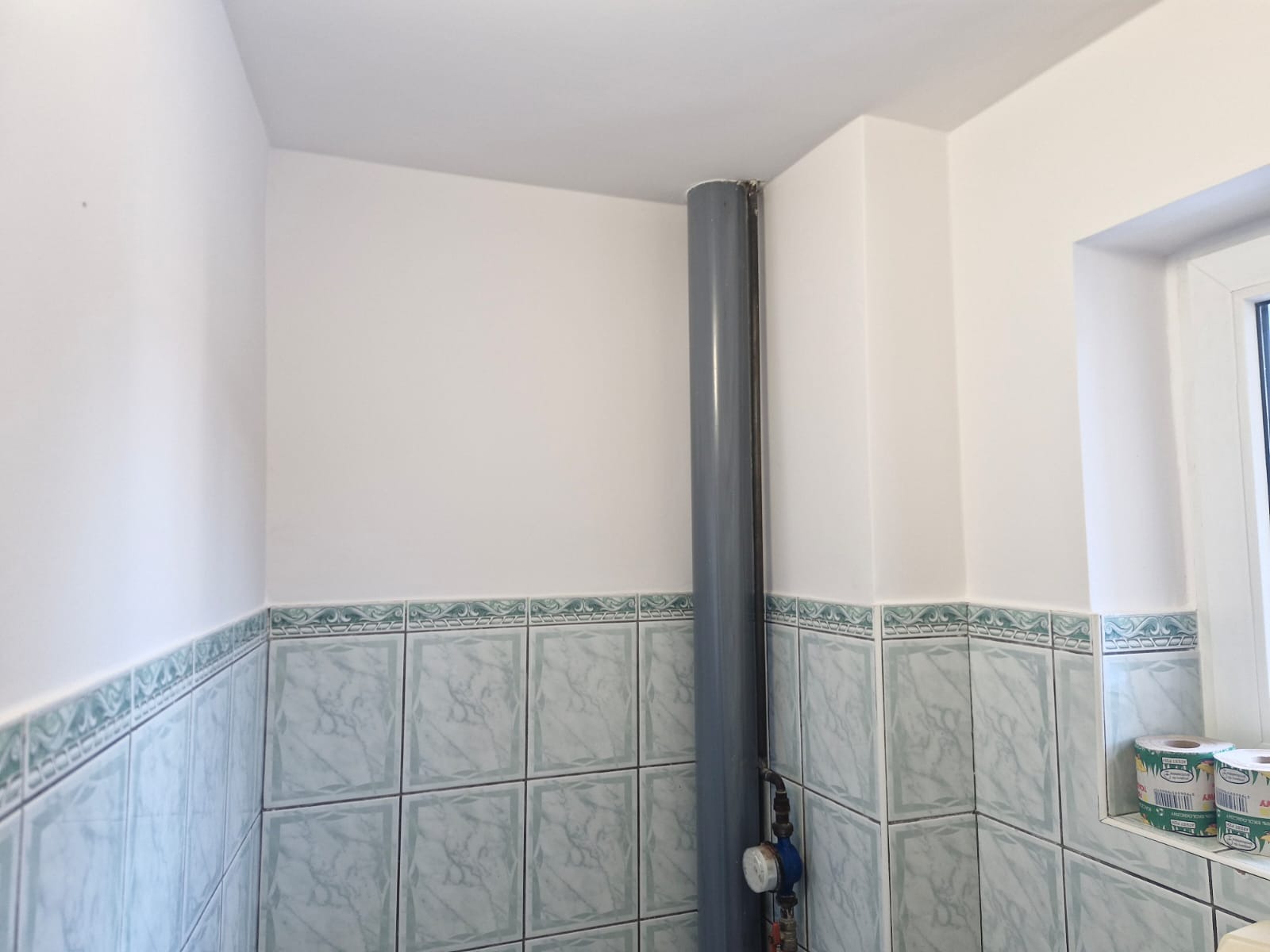 Szary pion kanalizacyjny w rogu łazienki z wykończonymi płytkami w tonacji błękitno-zielonej, widoczny manometr na rurze i fragment okna z rolkami papieru toaletowego na parapecie.