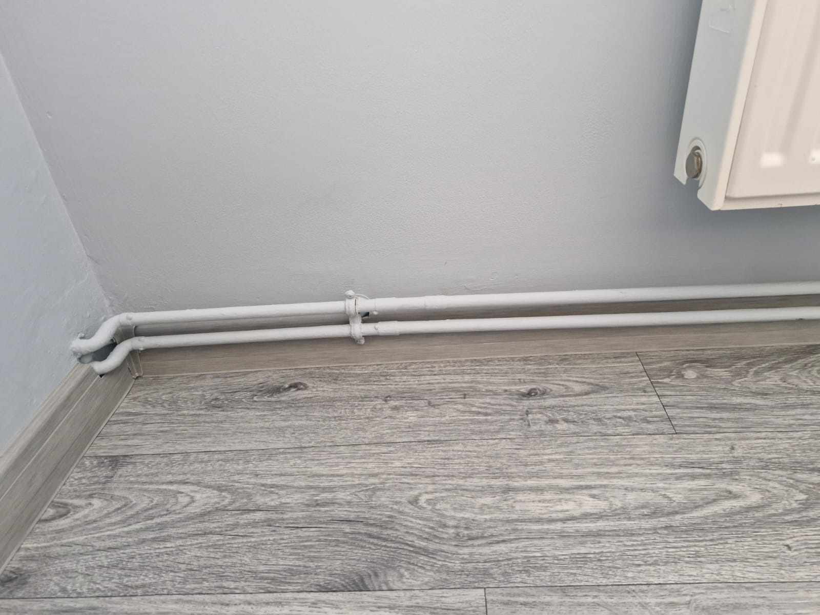 Dwie białe rury centralnego ogrzewania, pomalowane na biało, biegnące wzdłuż szarej podłogi panelowej i przy białej ścianie, widoczne obejmy mocujące.