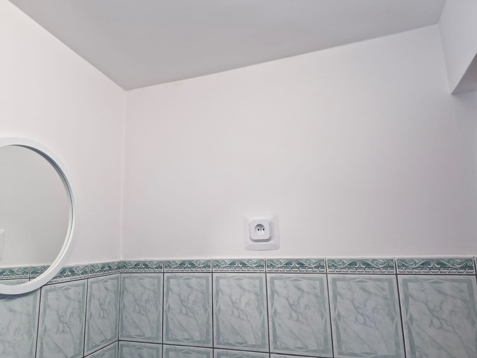 Biała ściana w łazience w Borzytuchom po malowaniu, z okrągłym lustrem i gniazdkiem elektrycznym nad pasem dekoracyjnych, marmurkowych płytek.