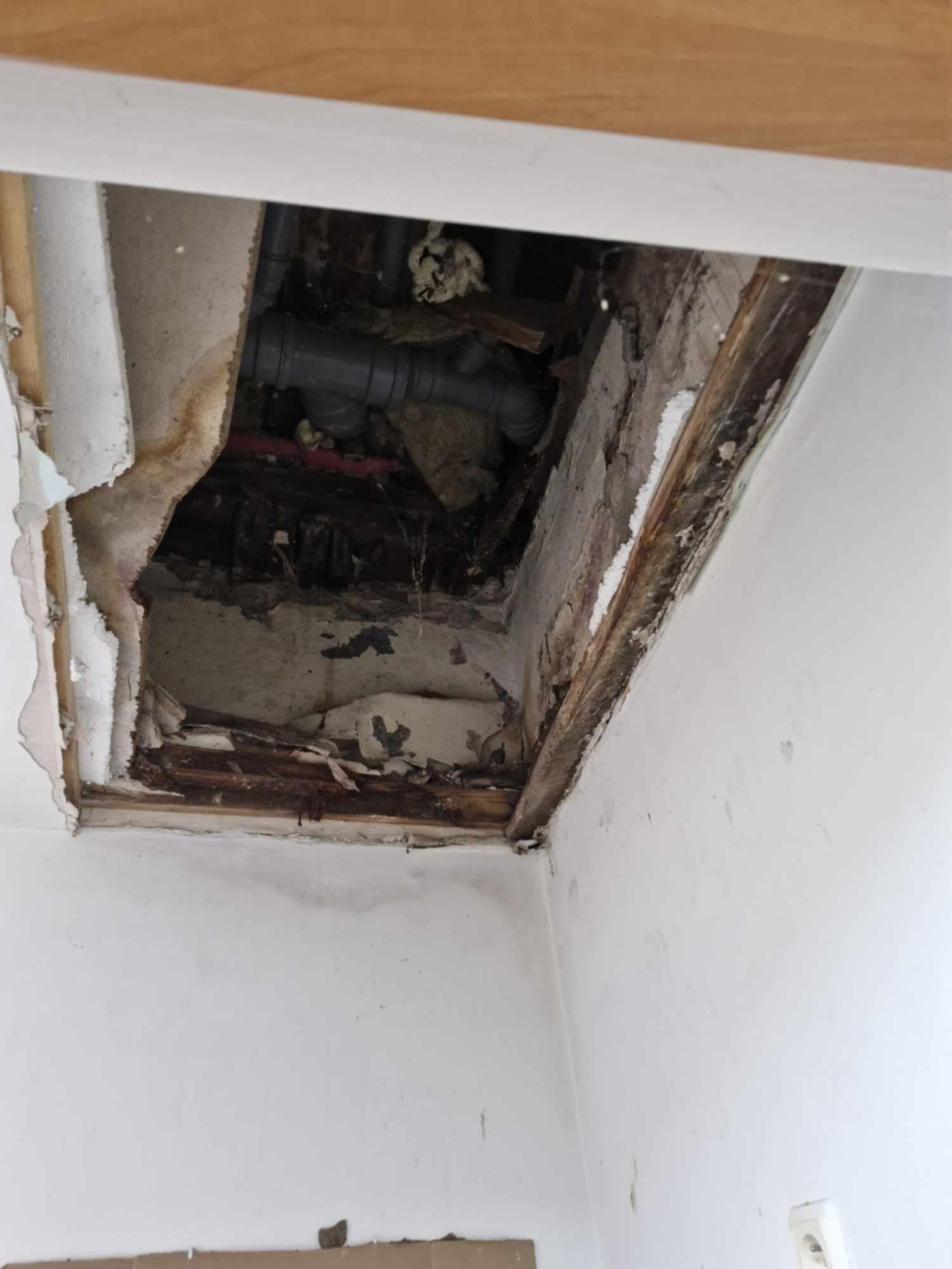 Otwór w suficie odsłaniający rury instalacyjne, widoczne uszkodzenia płyty kartonowo-gipsowej i drewnianej konstrukcji, fragment ściany z gniazdkiem elektrycznym w tle.