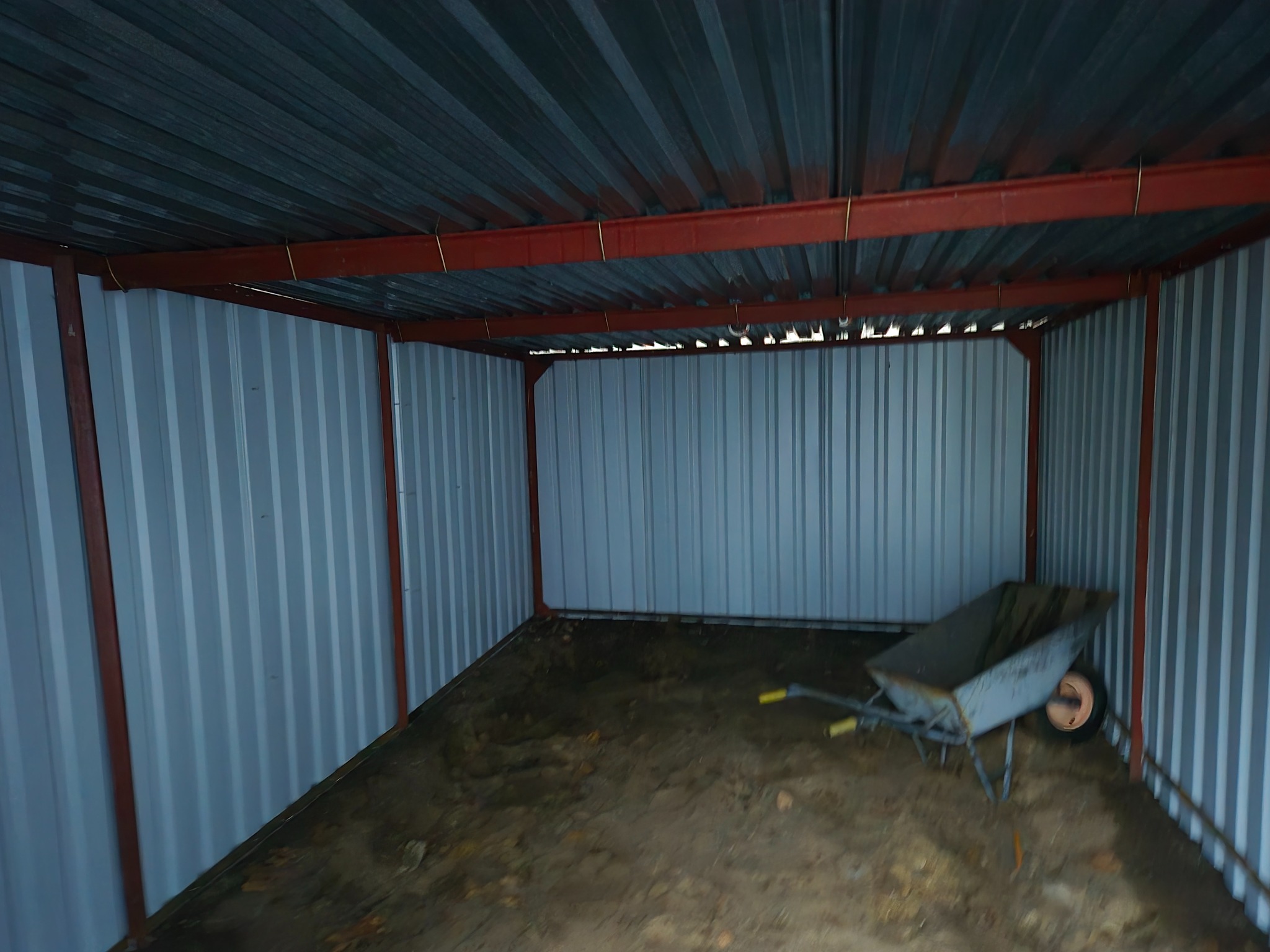 Wnętrze blaszanego garażu z czerwoną konstrukcją nośną dachu i brudną taczką po prawej stronie, widok od frontu.