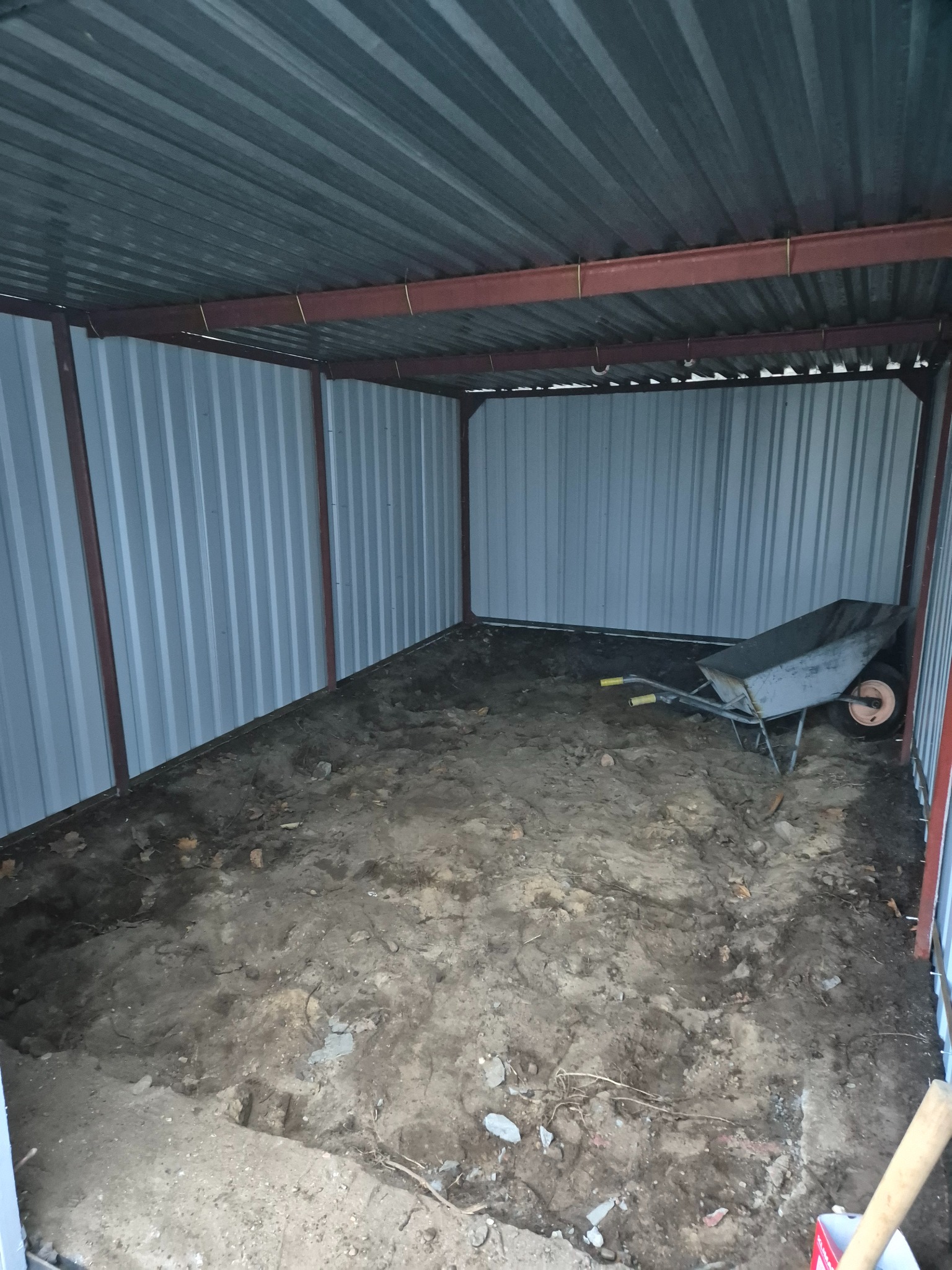 Wnętrze blaszanego garażu lub magazynu z nierównym, ziemistym podłożem i zaparkowaną taczką z żółtymi uchwytami.