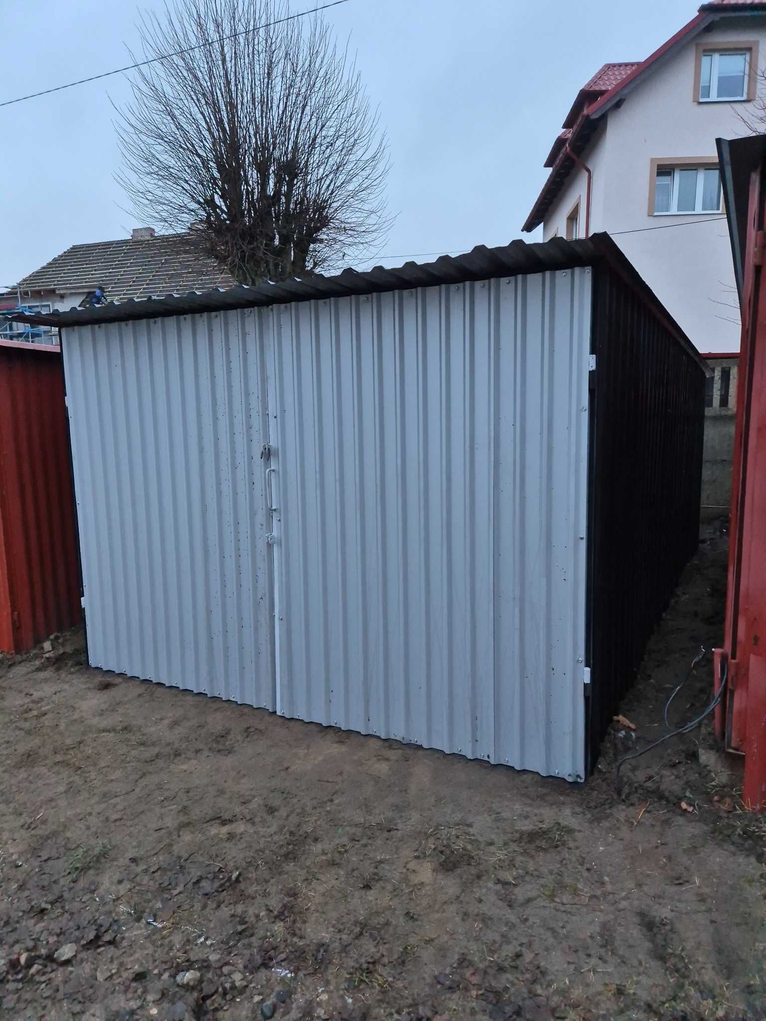 Nowo postawiony, blaszany garaż z ryflowanej blachy w kolorze jasnoszarym, z czarnymi bokami, stojący na błotnistym podłożu, z widocznym fragmentem domu w tle.