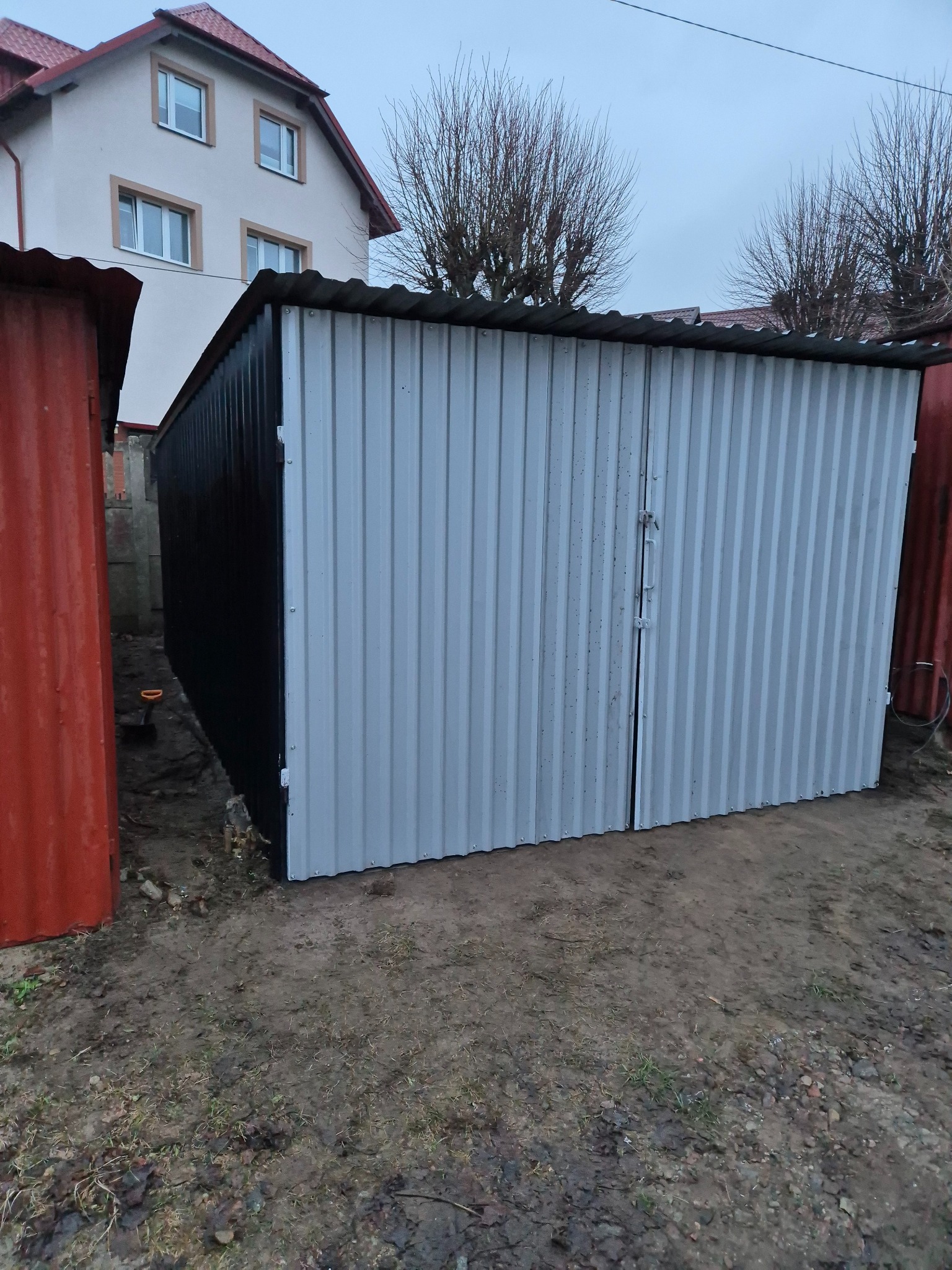 Metalowy garaż blaszany, pomalowany na czarno i biało, widoczny na tle domu jednorodzinnego i drzew bez liści.