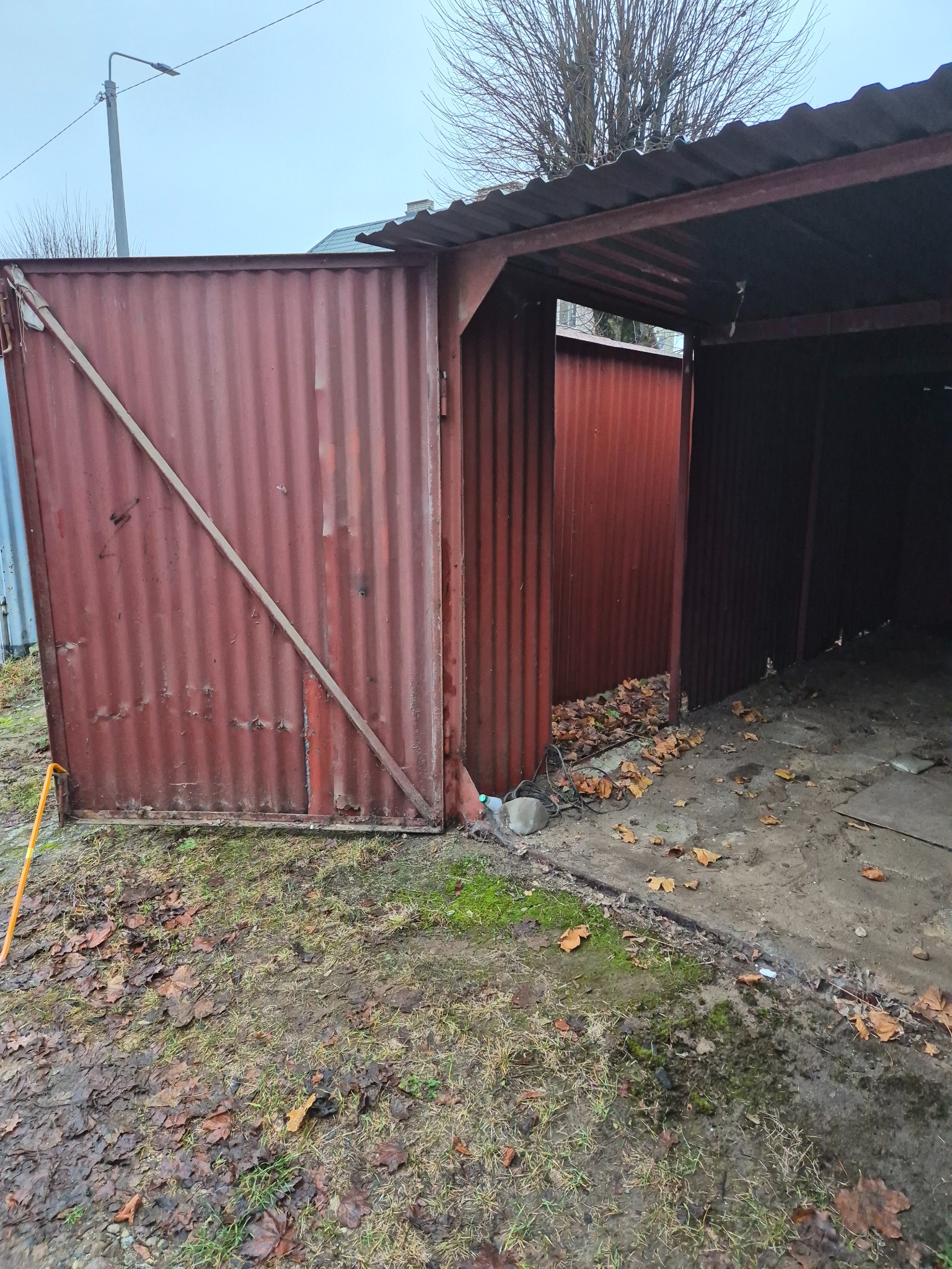 Otwarta, zardzewiała, blaszana wiata garażowa z widocznymi śladami korozji i brudu, otoczona błotnistym terenem z opadłymi liśćmi.