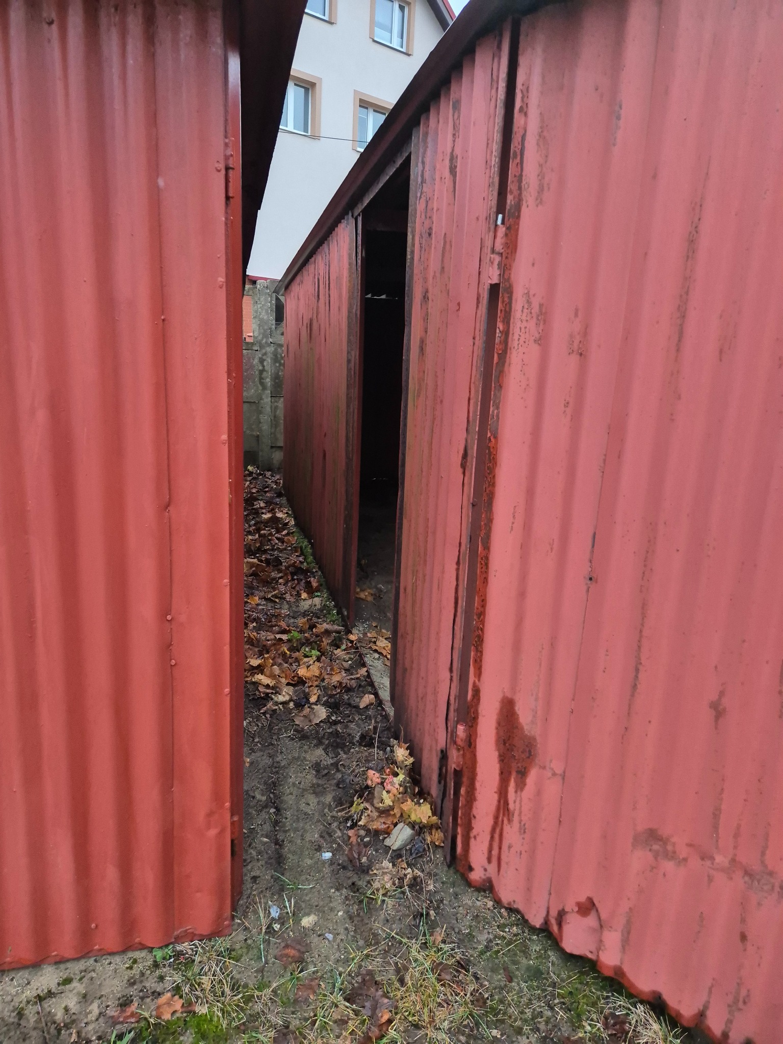 Czerwony, blaszany garaż z otwartymi drzwiami, widoczne liście na ziemi i fragment budynku w tle.