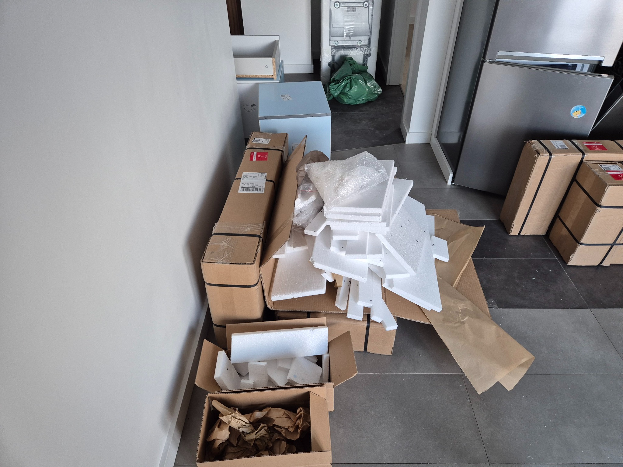Północno-zachodni kadr na kartony, styropianowe zabezpieczenia i papier pakowy po rozpakowaniu mebli, widoczne sprzęty AGD w tle.