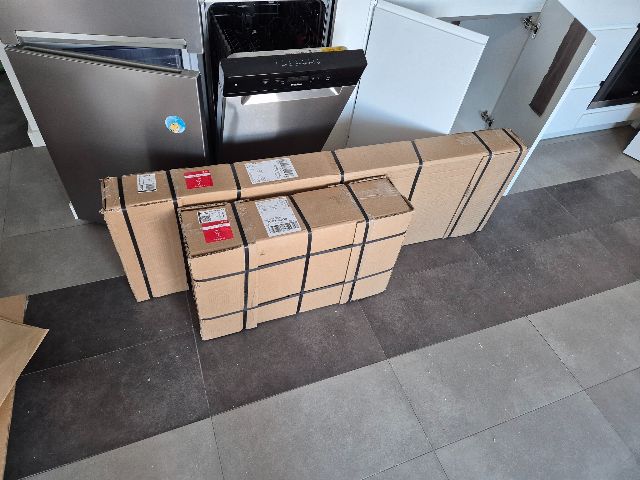 Paczki z nowymi meblami lub sprzętem AGD leżące na podłodze w kuchni, obok zmywarki i szafek kuchennych z otwartymi drzwiczkami.