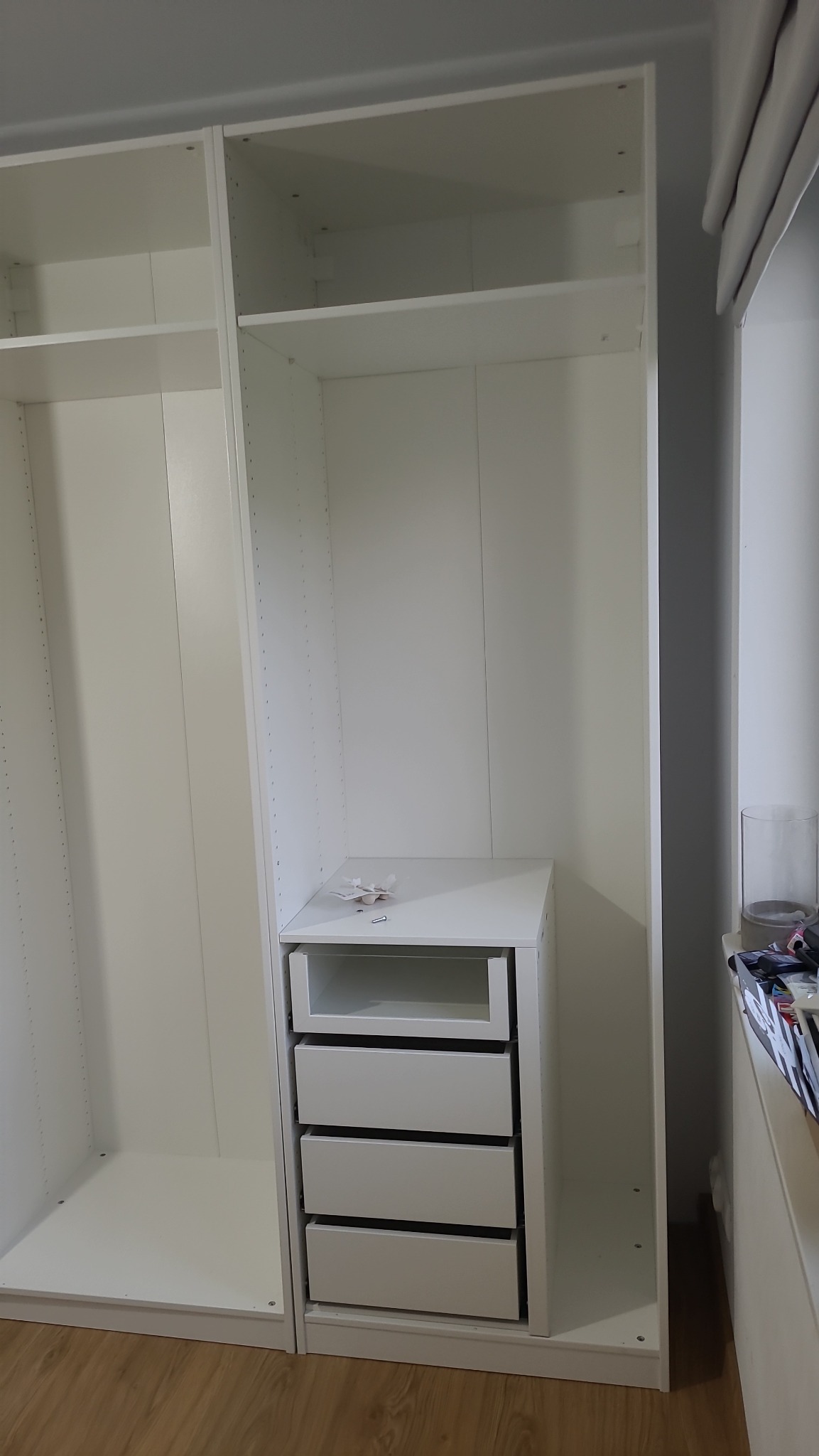 Biała szafa z szufladami w trakcie montażu, widoczne elementy mocujące na blacie, wnętrze szafy puste, podłoga drewniana.