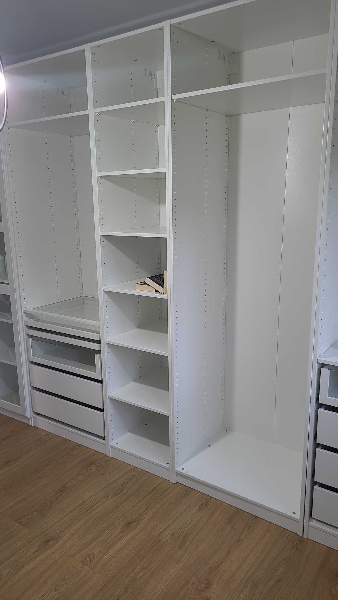 Biała, modułowa garderoba z półkami, szufladami i przestrzenią na wieszaki, z książkami na jednej z półek, na podłodze panele drewnopodobne.