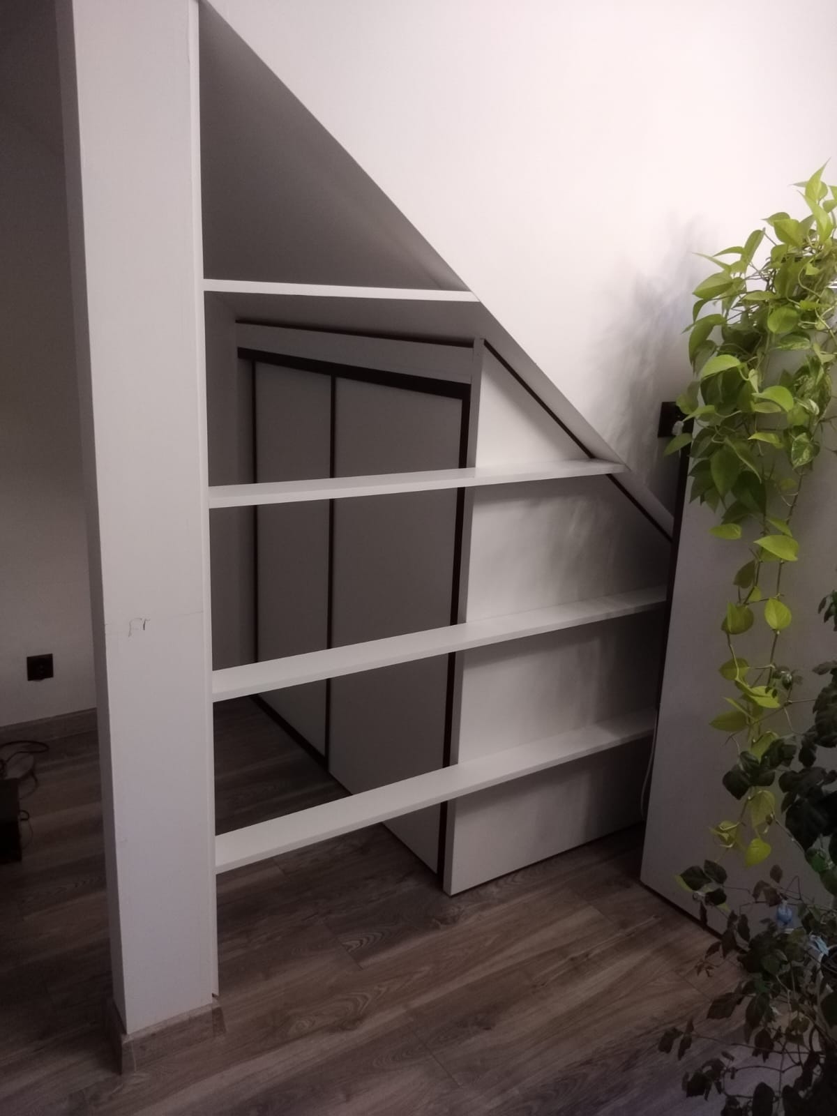 Biała, niestandardowa półka pod skosem schodów, z widocznymi drzwiami szafy w tle, uzupełniona zieloną rośliną doniczkową.