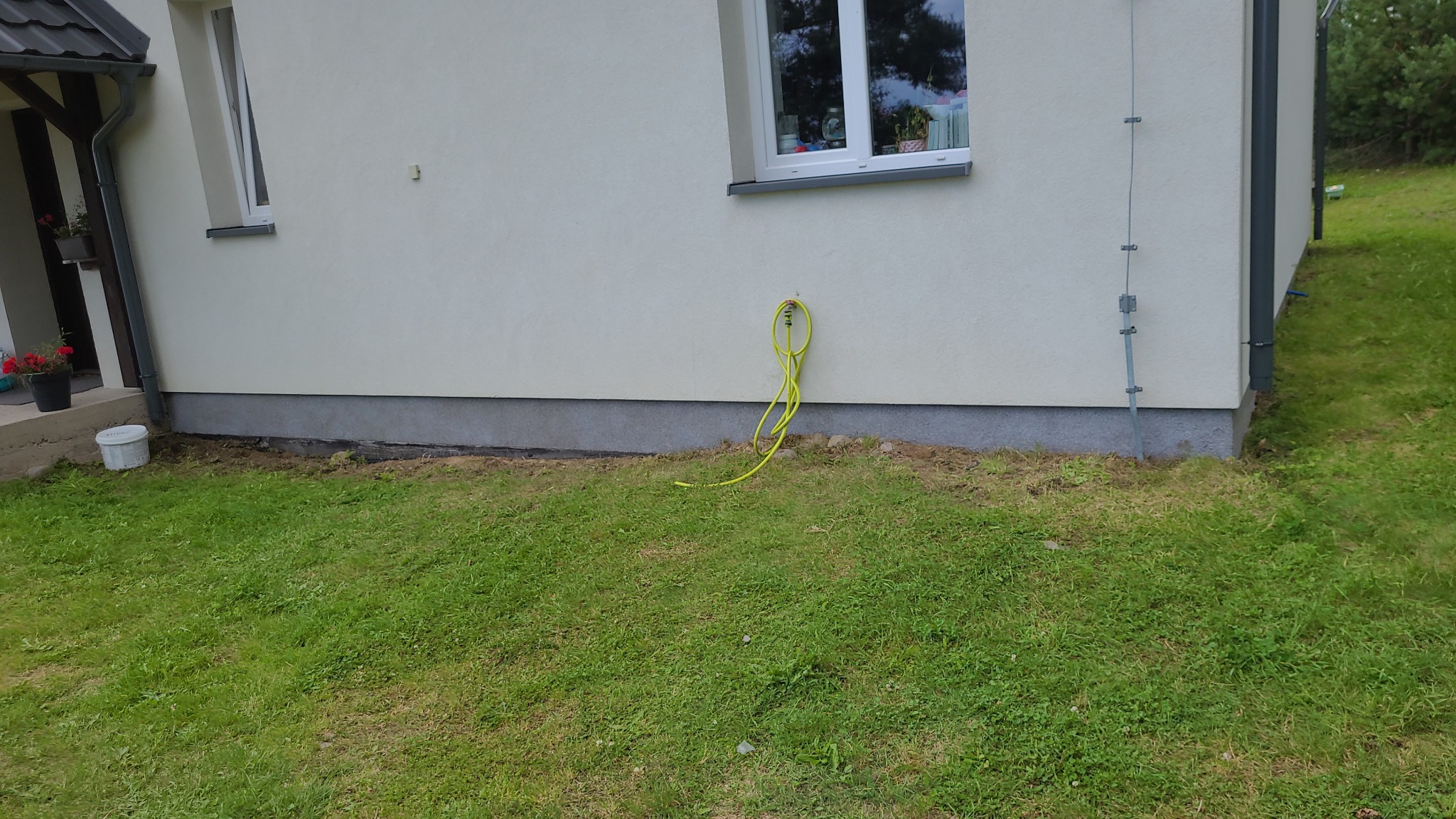 Fragment elewacji domu z widocznym żółtym wężem ogrodowym zwisającym ze ściany, trawnik z ziemią przy fundamencie, okno z roślinami w tle.