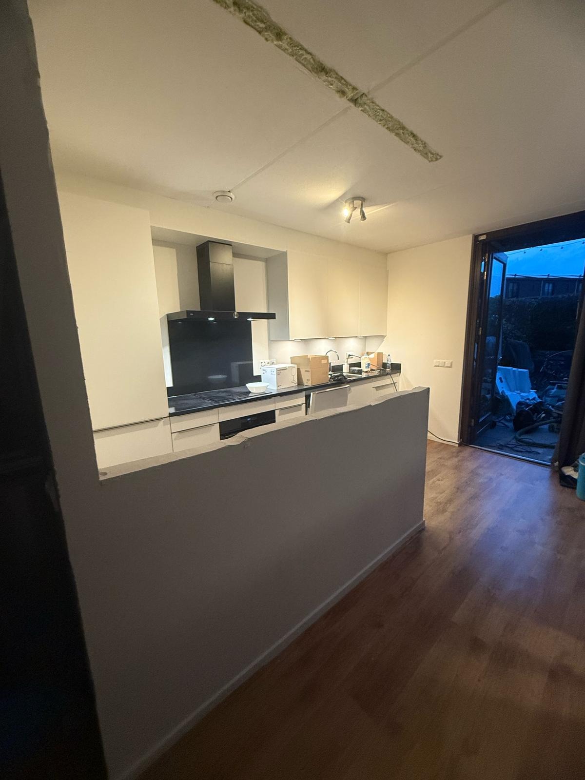 Jasna kuchnia z wyspą w trakcie remontu: widoczne niedokończone sufity i otwarte drzwi na zewnątrz. Podłoga z paneli, białe szafki, czarny okap. Ujęcie z boku.