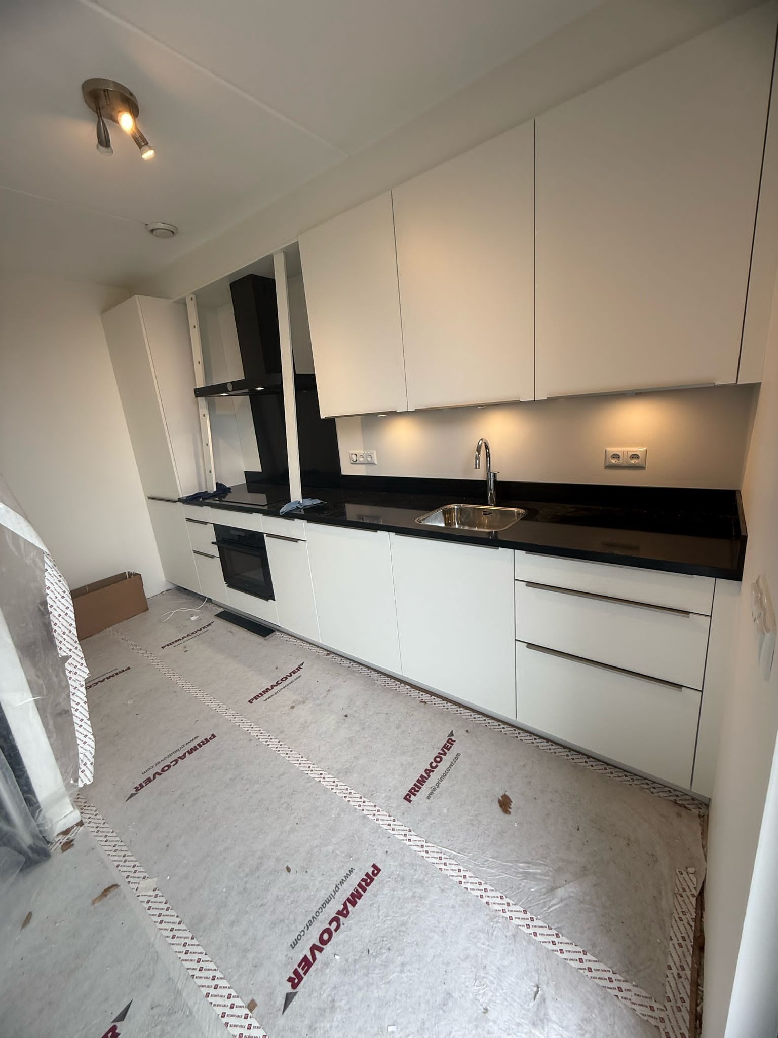 Nowoczesna kuchnia z białymi szafkami i czarnym blatem, oświetlona punktowo. Podłoga zabezpieczona folią ochronną. Minimalistyczny design.