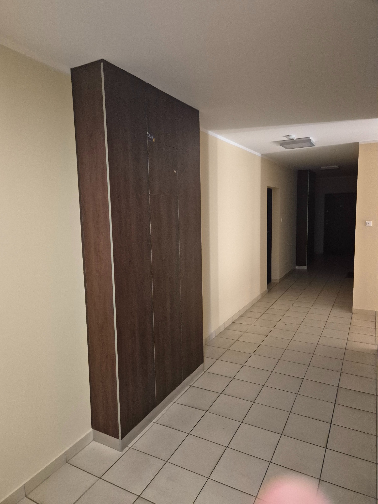 Wysoka, ciemnobrązowa szafa wnękowa z metalowymi listwami, zamontowana w korytarzu z jasnymi ścianami i kafelkami. Widok na długi korytarz z drzwiami.