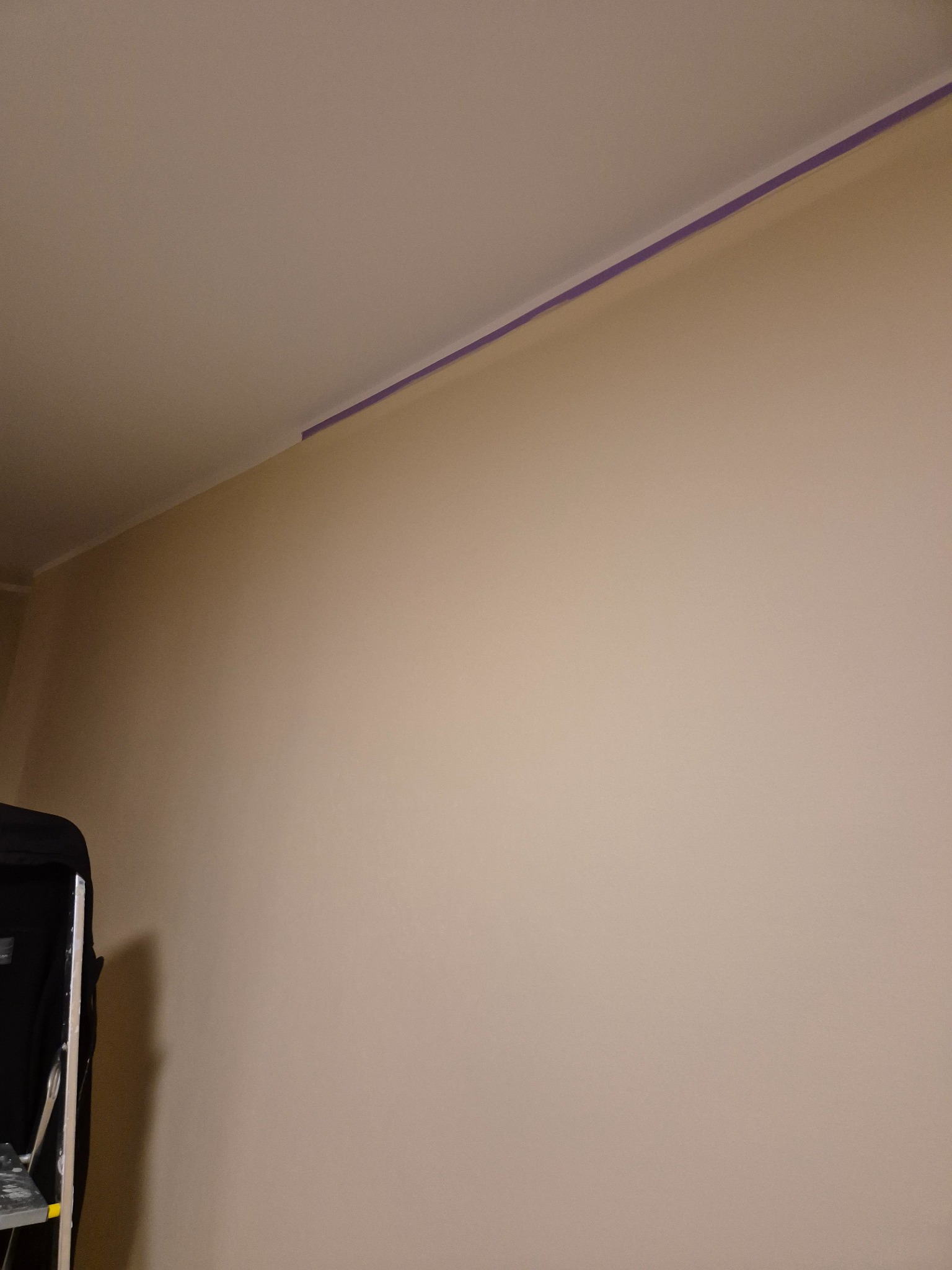 Ściana w trakcie malowania, zabezpieczona taśmą malarską przy suficie. Widoczny fragment drabiny malarskiej. Beżowa farba na ścianie.