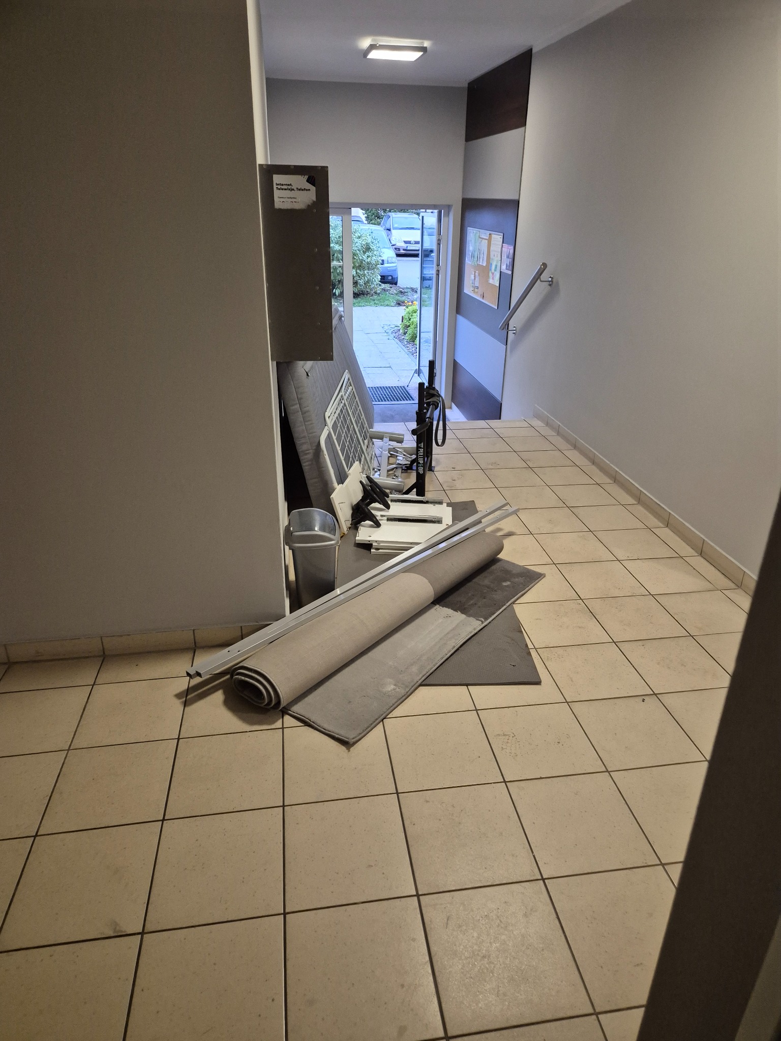 Korytarz w bloku z przygotowanymi materiałami do remontu: zwinięty dywan, listwy przypodłogowe, drabina do ćwiczeń. Widok na zewnątrz przez otwarte drzwi.