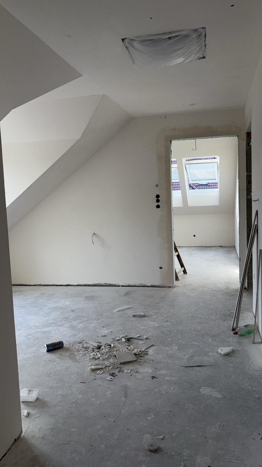 Wnętrze poddasza w trakcie remontu: białe ściany, surowy beton na podłodze z resztkami gruzu, okno dachowe. Widoczne przygotowania do dalszych prac wykończeniowych.