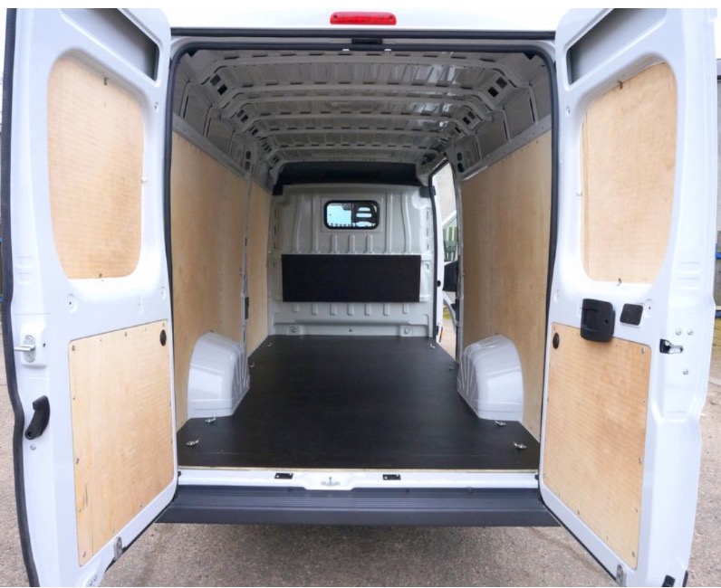 Wnętrze białego busa wyłożone sklejką na ścianach i czarną podłogą, otwarte tylne drzwi.