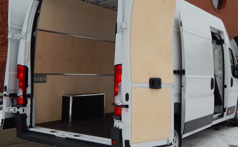 Wnętrze białego furgonetki z otwartymi tylnymi drzwiami, wyłożone sklejką i wyposażone w metalowe uchwyty do mocowania ładunku.