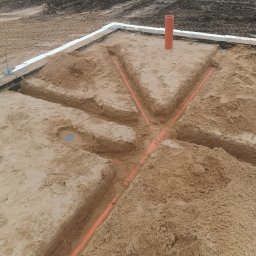 HYDRO Instal Mateusz Aseńczuk - Przygotowanie podłoża pod ogrzewanie podłogowe: wykopane rowki w piasku z ułożonymi rurami grzewczymi, widoczna izolacja fundamentów i pionowa rura wentylacyjna.